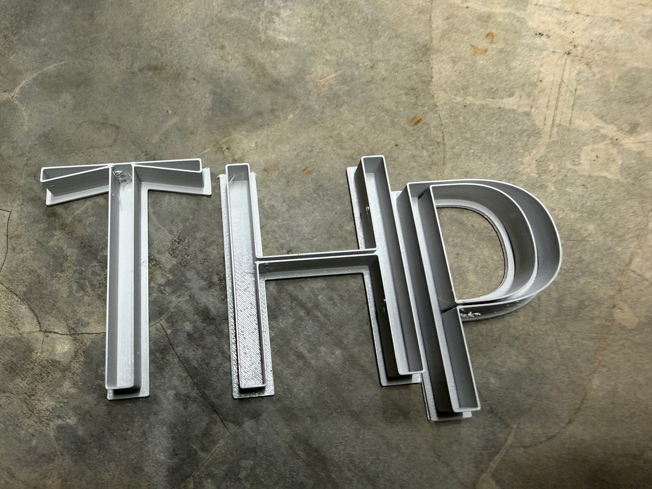 ตัวอักษร THP สีเทาวางบนพื้น แสดงโครงสร้างตัวอักษรแบบกลวง